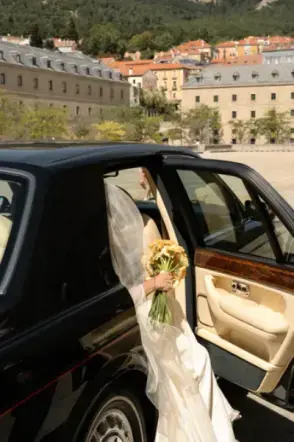 Novia saliendo de un coche clásico negro con un ramo de flores amarillas en la mano y velo largo.