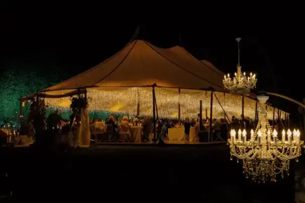 Banquete de boda nocturno bajo una gran carpa iluminada con luces cálidas y lámparas de araña.